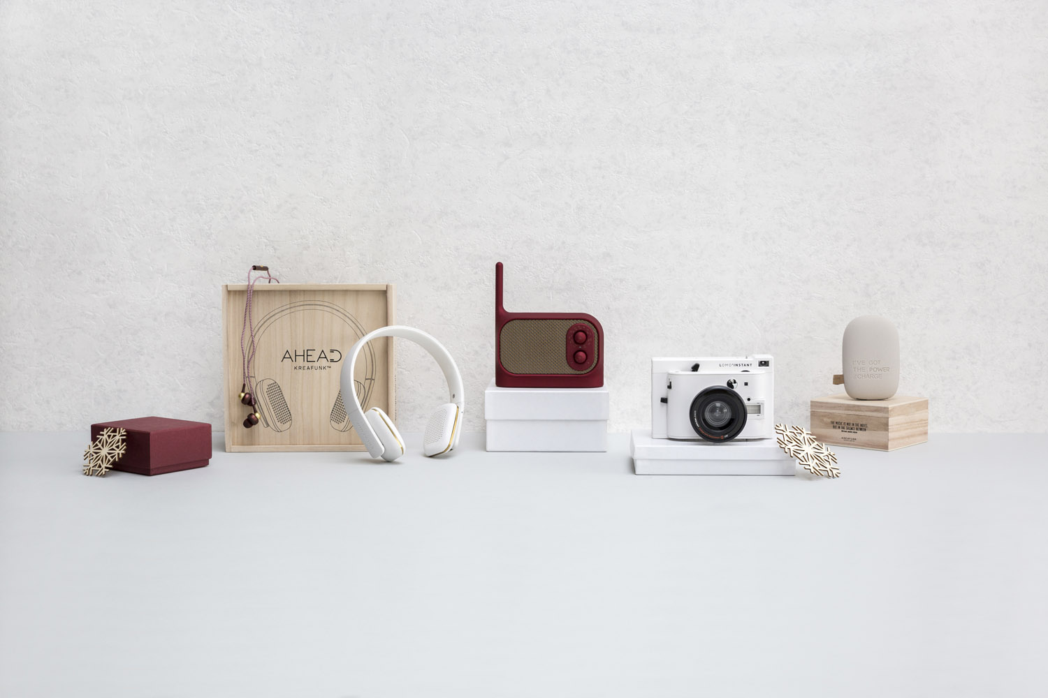 Various tech gadgets and accessories, including headphones, a portable speaker, a camera, and an oval-shaped device, are neatly arranged on white and wooden boxes against a light grey background.