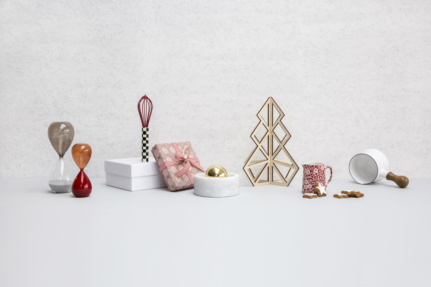 A collection of objects on a light surface: two hourglasses, small boxes, a gift wrapped in pink paper, a whisk, a marble container with a gold sphere, a wooden tree decoration, a mug, scattered biscuits, and an upturned bowl.