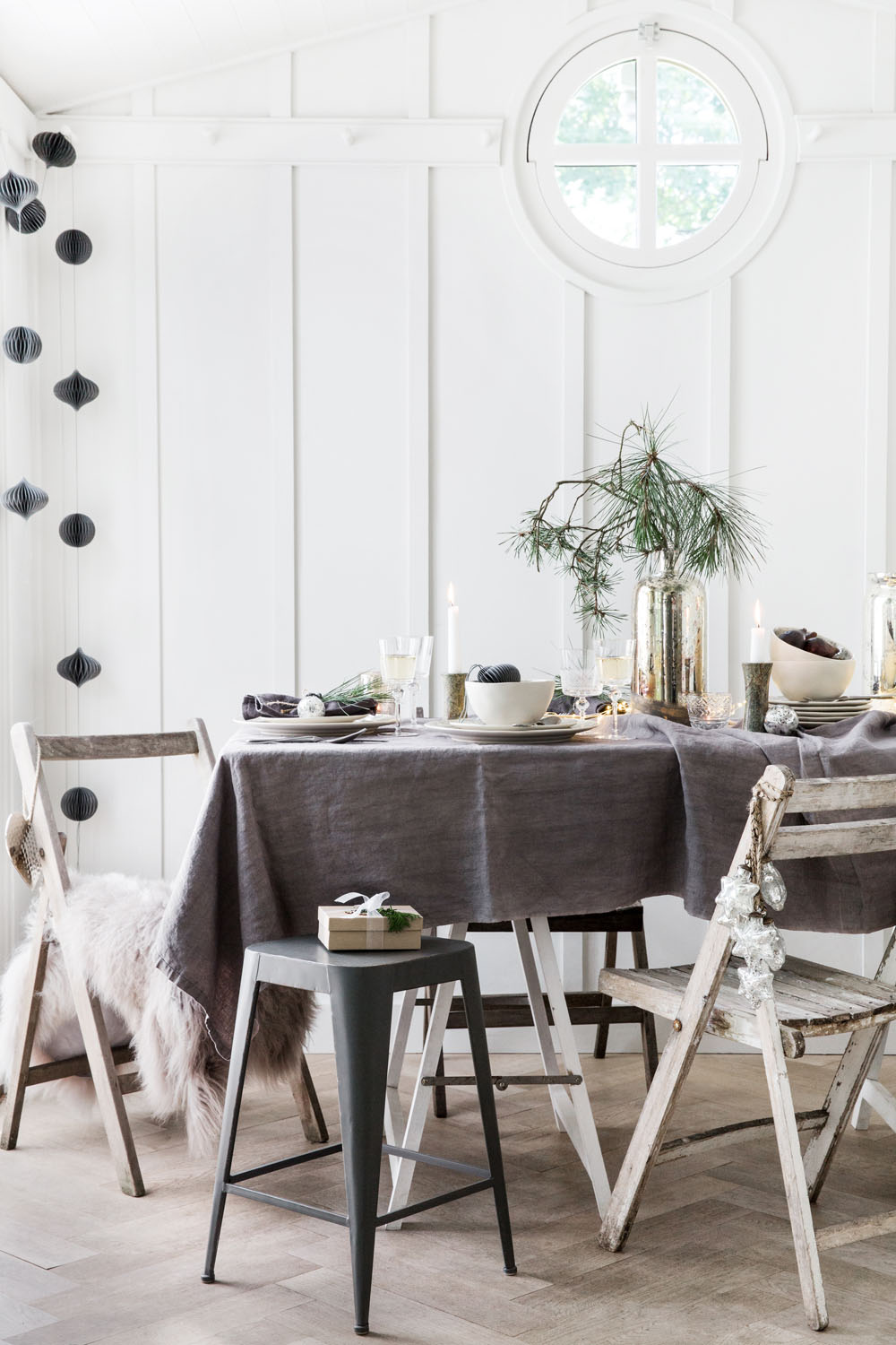 A small dining table covered with a grey tablecloth is set with crockery and candles. Three rustic wooden chairs and a black stool surround the table. A small window and white panelled walls are in the background.