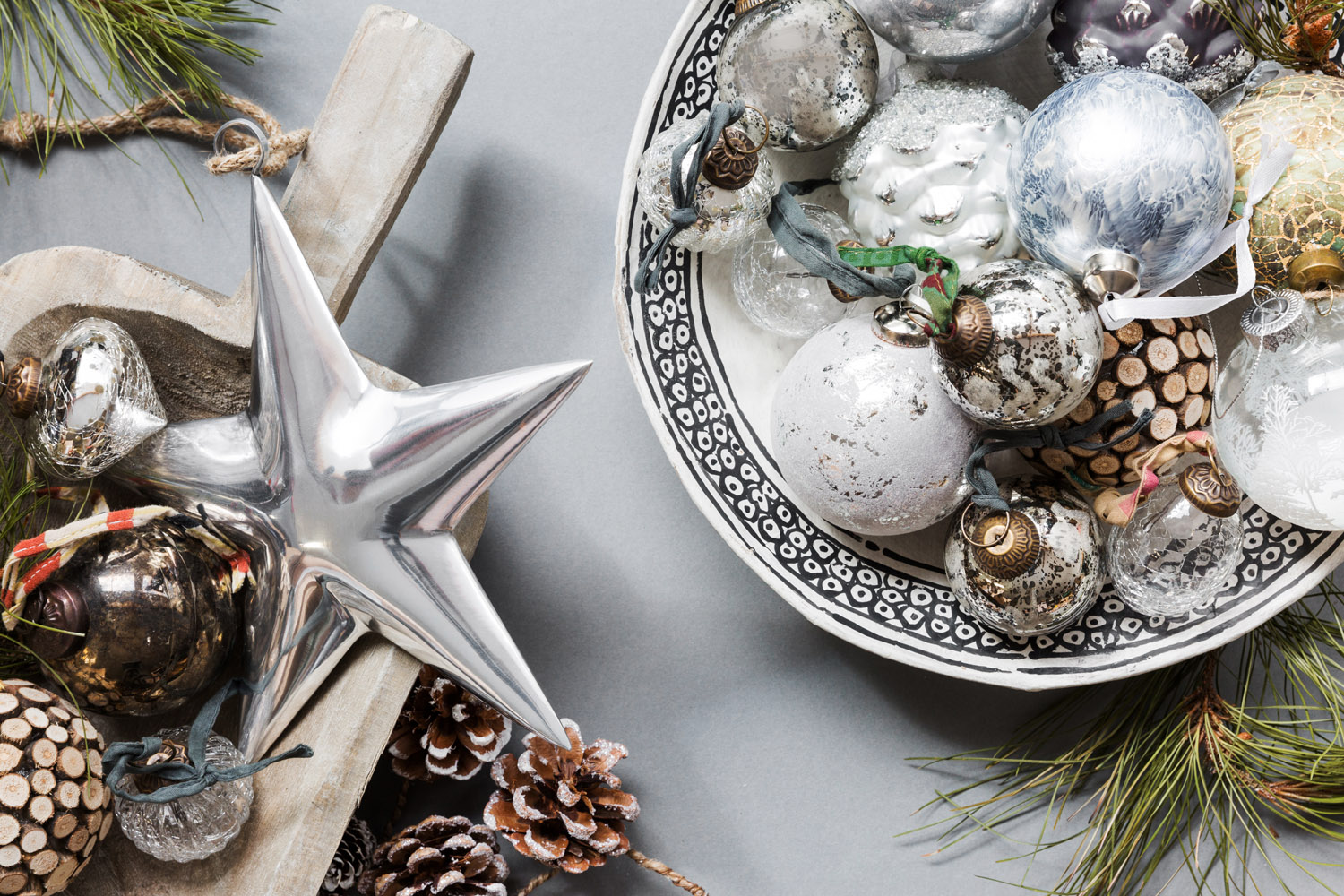 A wooden tray with a large silver star, pine cones, and baubles sits next to a patterned bowl filled with assorted Christmas tree decorations and wooden slices, surrounded by pine branches on a grey surface.