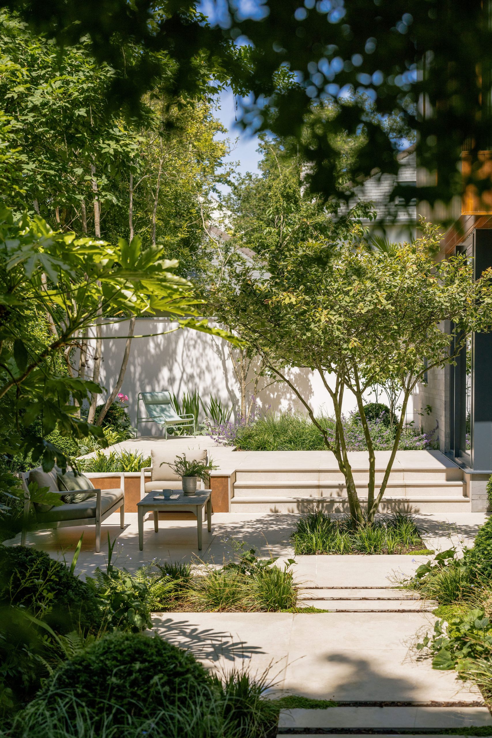 A modern garden with stone pathways, green plants, and trees. There are wooden outdoor chairs and a small table on a patio, with sunlight filtering through the foliage. The setting appears quiet and inviting.