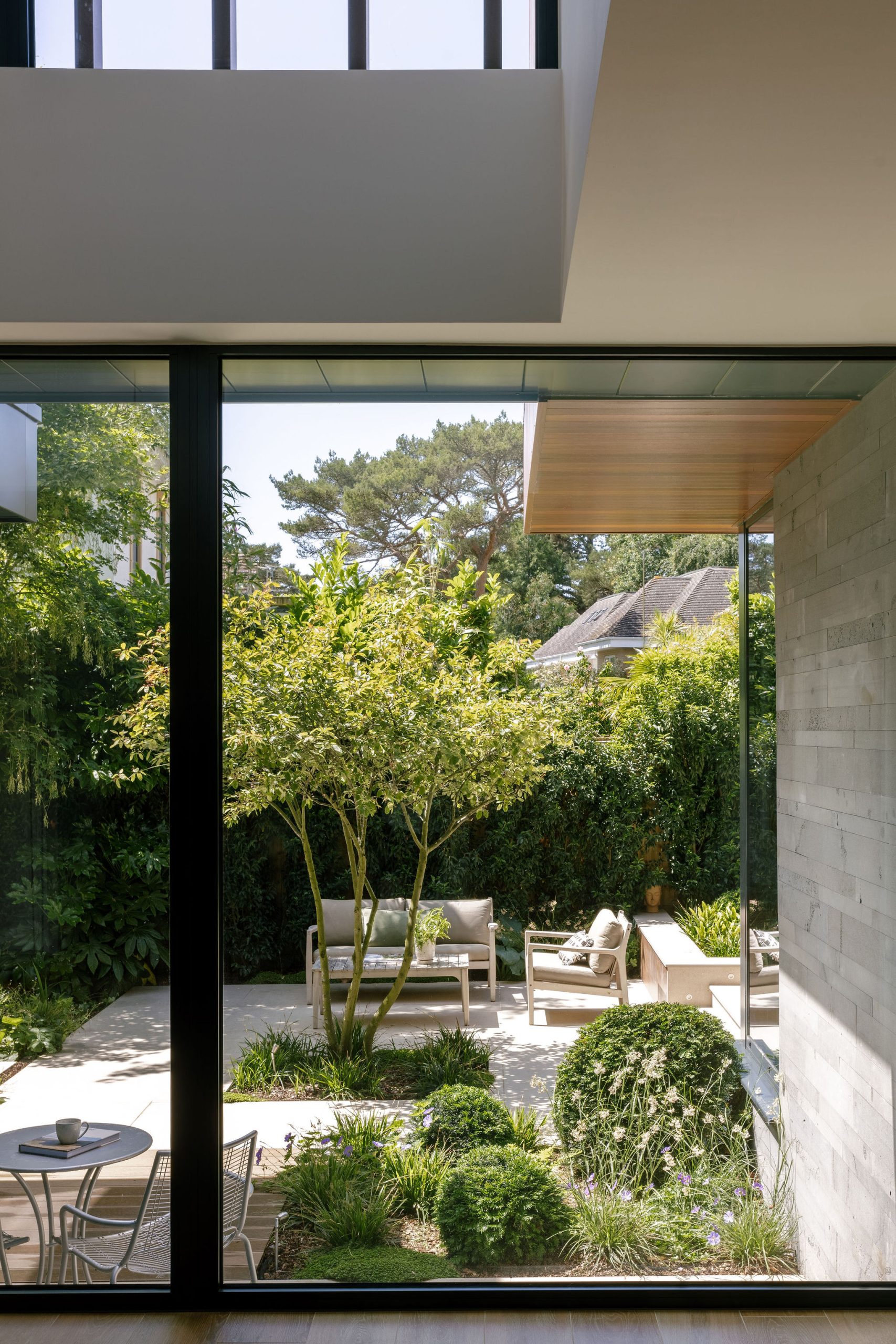 View through a large glass window showing a modern patio with outdoor seating, small tables, neatly trimmed bushes, and trees, surrounded by greenery and a garden, with sunlight filtering through.