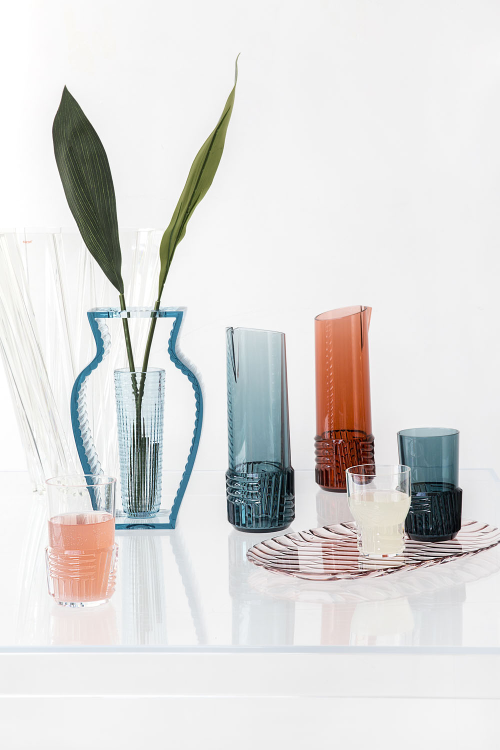 A clear table displays colourful glassware, including two jugs (one blue, one orange), matching glasses, a blue-edged vase with green leaves, and striped serviettes, all arranged against a white background.