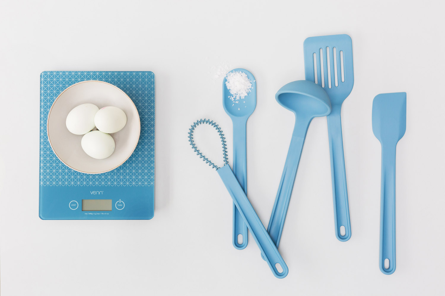 A digital kitchen scale with a bowl of three eggs sits next to five blue kitchen utensils, including a spoon with salt, a whisk, a ladle, a slotted fish slice, and a spatula, arranged on a white surface.