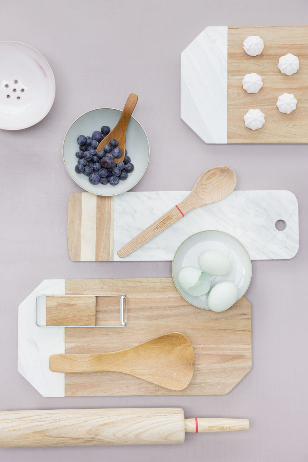 A flat lay of wooden and marble chopping boards, a peeler, a wooden spoon, a rolling pin, a plate of blueberries, a small bowl with eggs, another bowl with meringues, and a pink dish on a light surface.