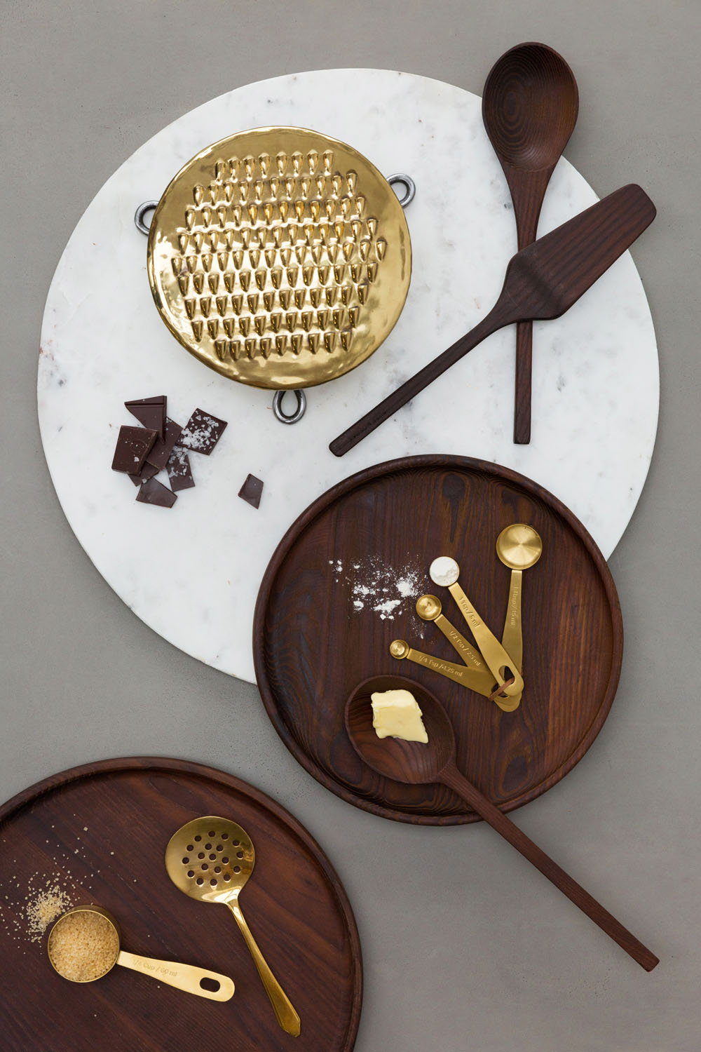 A flat lay of dark wooden utensils, gold measuring spoons, a brass grater, a slotted spoon, broken chocolate pieces, and a pat of butter on marble and wooden boards with some scattered flour and sugar.