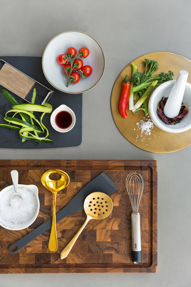 Overhead view of kitchen utensils, a whisk, slotted spoon, and measuring spoon on a wooden board, with bowls of salt, cherry tomatoes, a red pepper, broccolini, a mortar and pestle, and sliced cucumber on a grey surface.