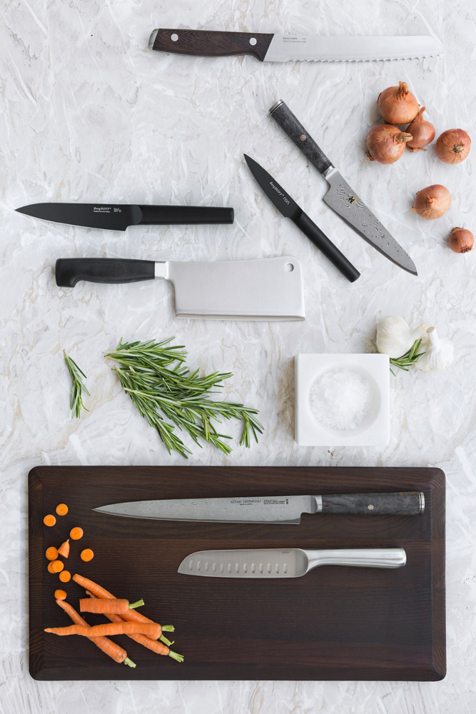 A selection of knives, herbs, and vegetables are arranged on a marble surface. A chopping board holds a chef’s knife, a santoku knife, and sliced carrots with whole carrots beside them. Garlic, onions, rosemary, and a bowl of salt are nearby.