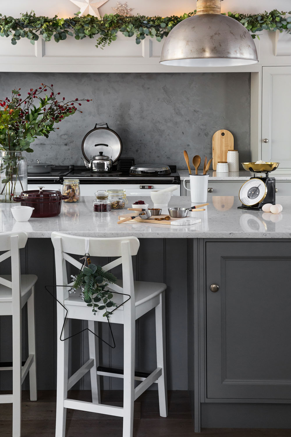 A modern kitchen with white and grey cupboards, a marble worktop, wooden utensils, and a metal hanging light. Festive greenery and a star decoration adorn the space, with various kitchen items and mugs on the island.