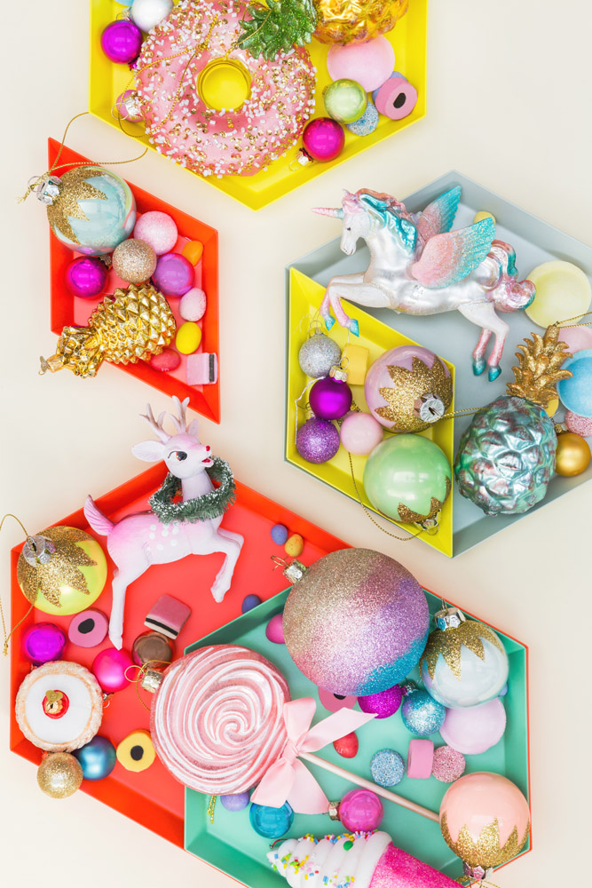 Colourful trays filled with decorative Christmas ornaments, including a unicorn, a pink deer, a doughnut, a lollipop, baubles, and novelty shapes, arranged on a cream background.
