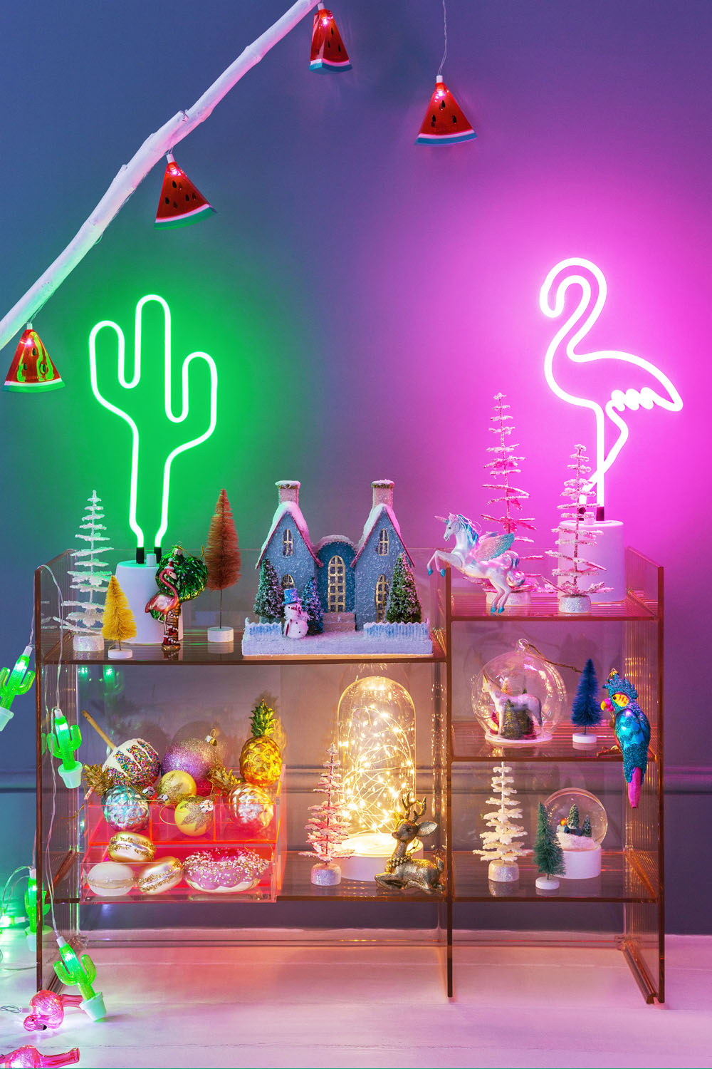 A shelving unit displays mini Christmas trees, baubles, and holiday figurines. Above are neon cactus and flamingo lights. Watermelon fairy lights hang on the wall, and the scene is lit with colourful glowing lights.