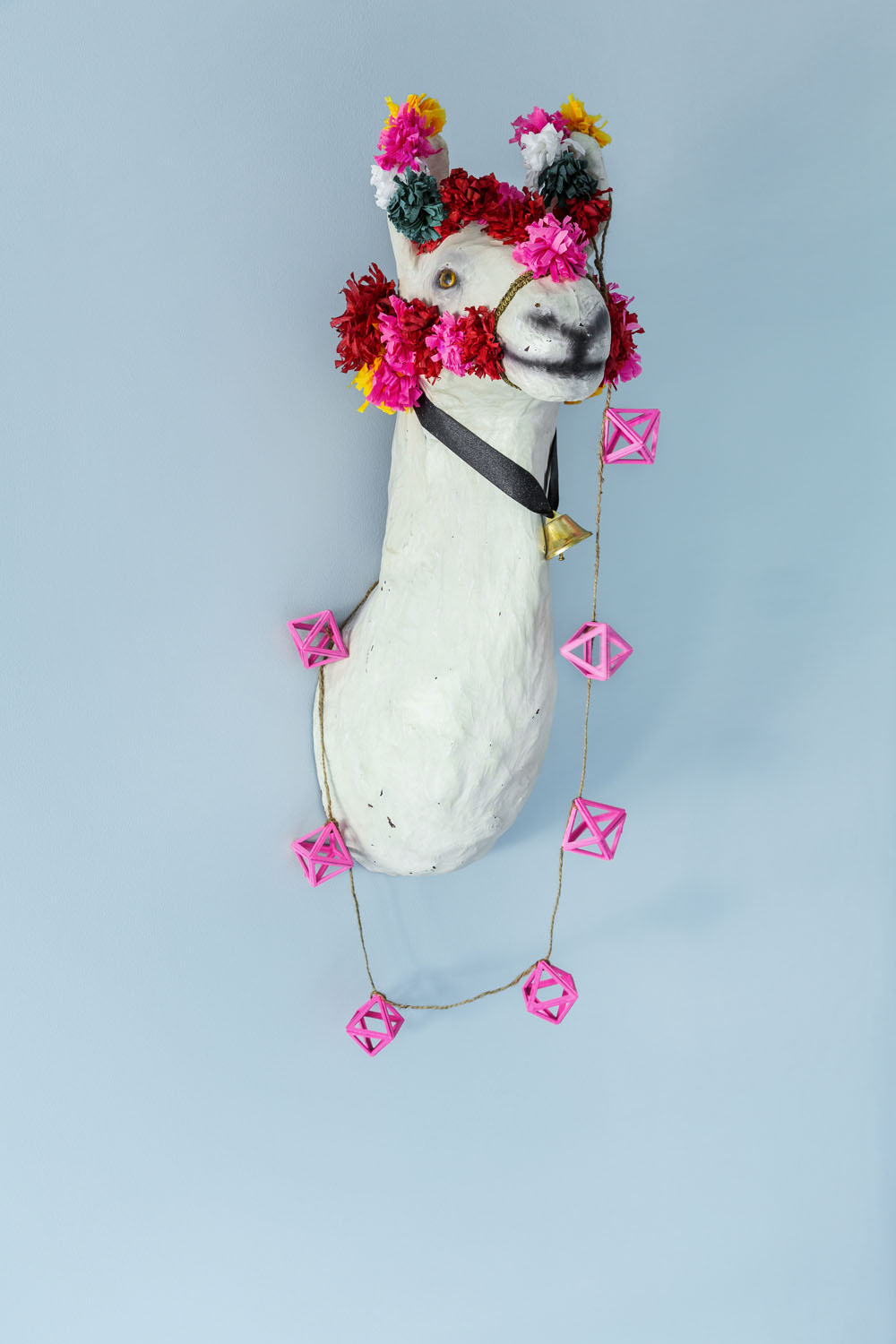 A white llama head sculpture is mounted on a light blue wall, decorated with colourful artificial flowers and a string of pink geometric ornaments hanging around its neck.