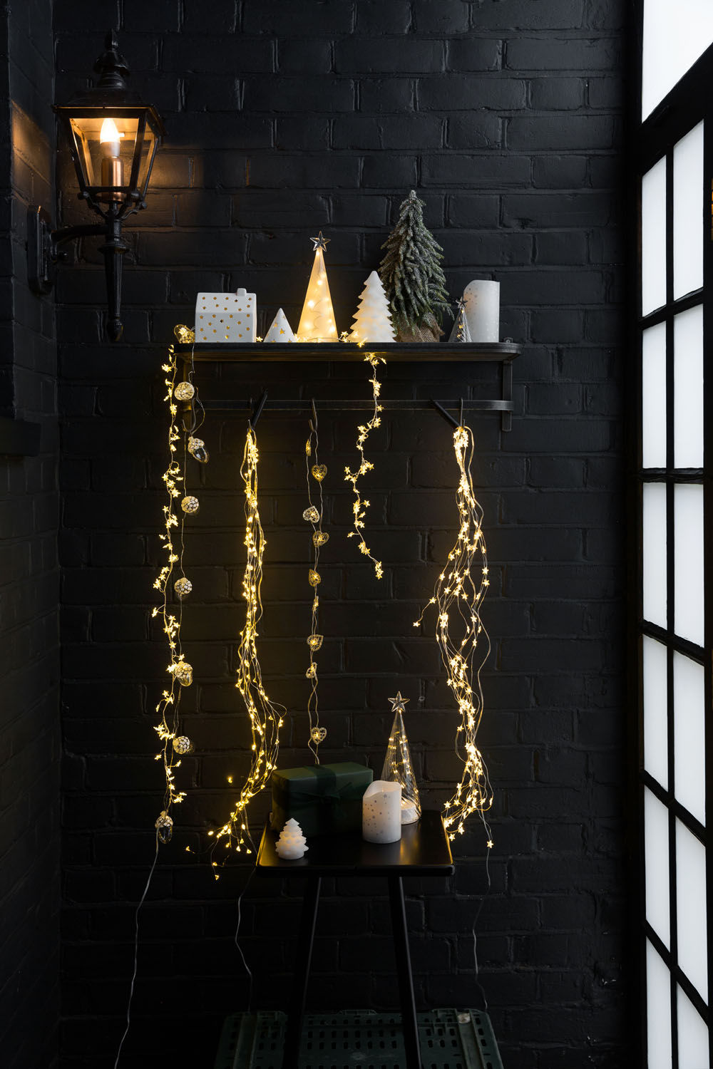 A black brick wall with a shelf holding small decorative trees, candles, and presents. Hanging from the shelf are strands of warm yellow fairy lights. A lantern is mounted on the wall next to a window with frosted glass panes.