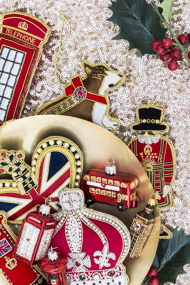 A collection of British-themed Christmas decorations, including a crown, red phone box, corgi, London bus, British flag, and a royal guard, arranged on a gold plate with holly leaves and berries on sequinned fabric.