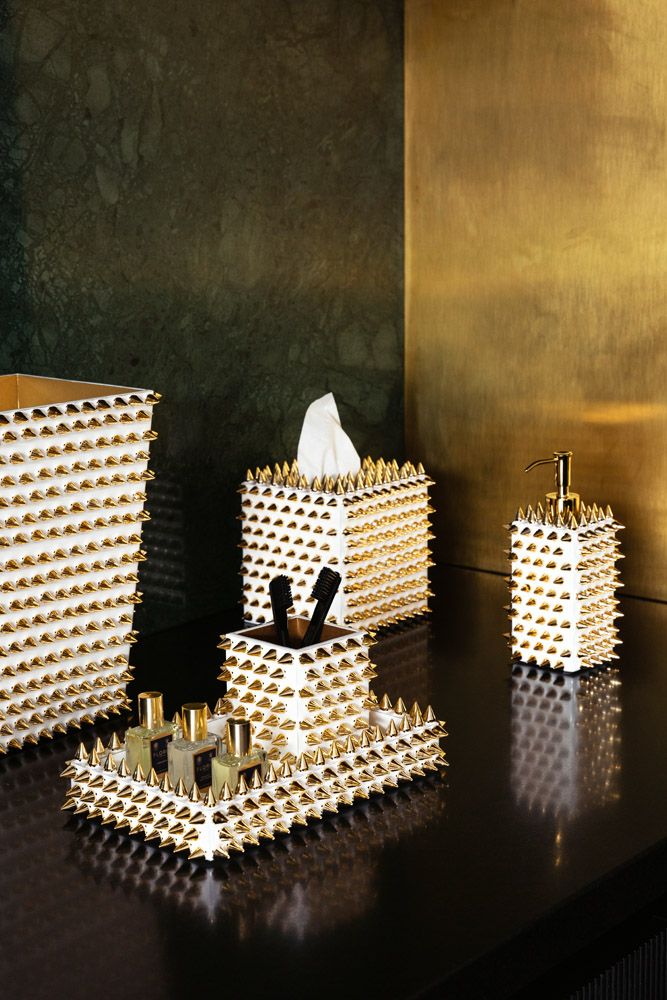 A black worktop displays white bathroom accessories with gold cone-shaped studs, including a tissue box, a tray holding bottles, a soap dispenser, and a waste bin. The background features dark green and gold surfaces.