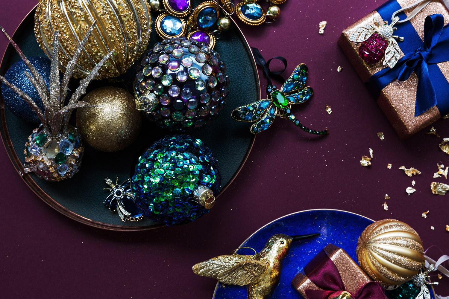 A flat lay of sequinned and glittery Christmas baubles, brooches, and wrapped presents with ribbons on a dark purple surface. Decorative balls, a dragonfly brooch, and a hummingbird ornament are featured.