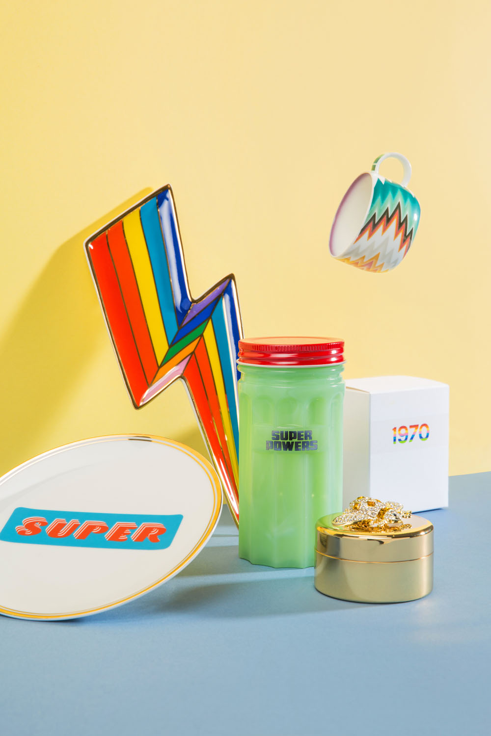 A colourful photo displays a rainbow lightning bolt, a green jar labelled Super Powers, a floating zigzag-patterned mug, a gold tin with jewellery, a white 1970 box, and a plate with SUPER text on a blue and yellow background.