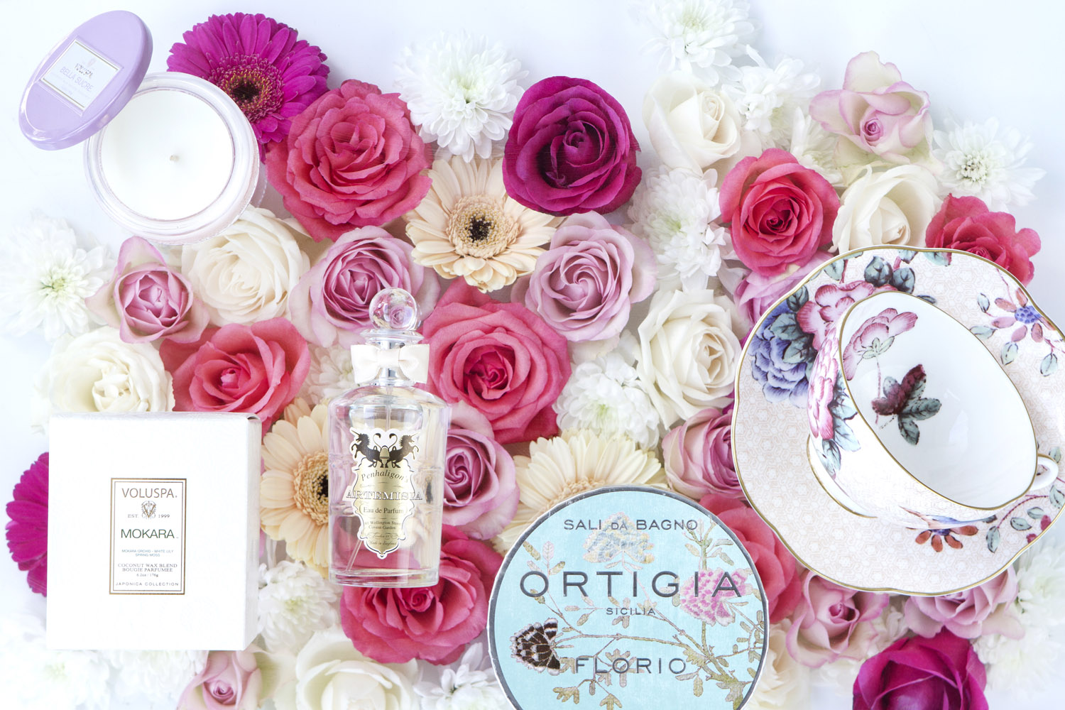 A flat lay of various pink and white flowers, a candle, a perfume bottle, a tin of bath salts, a small box, a soap container, and a decorative teacup and saucer with a butterfly design.