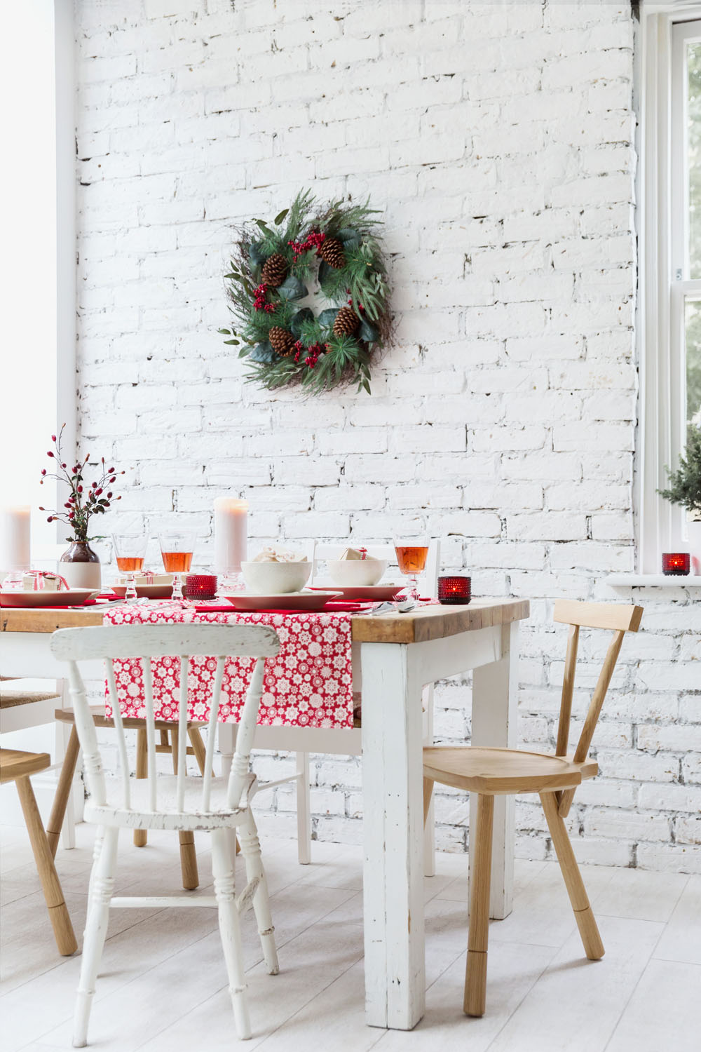 A white wooden dining table with mismatched chairs is set with dishes and glasses. A red and white tablecloth covers part of the table. Candles and a wreath with pine cones decorate the white brick wall and window.