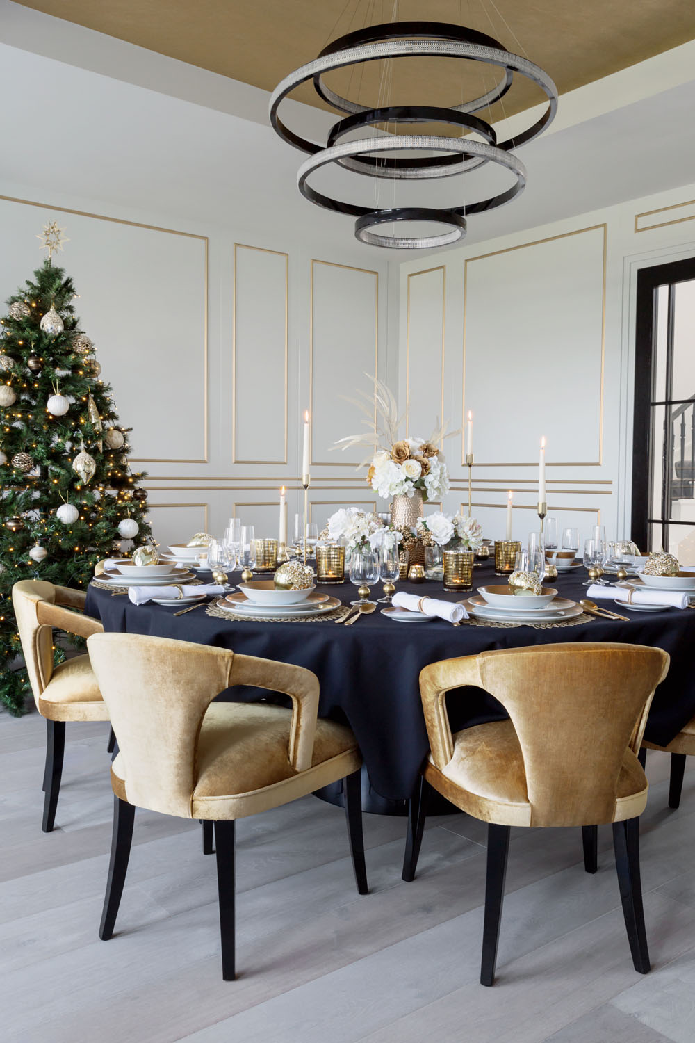 A round dining table with a black tablecloth is set for a formal meal with gold and white decorations, surrounded by six gold velvet chairs. A decorated Christmas tree stands in the corner of an elegant room.