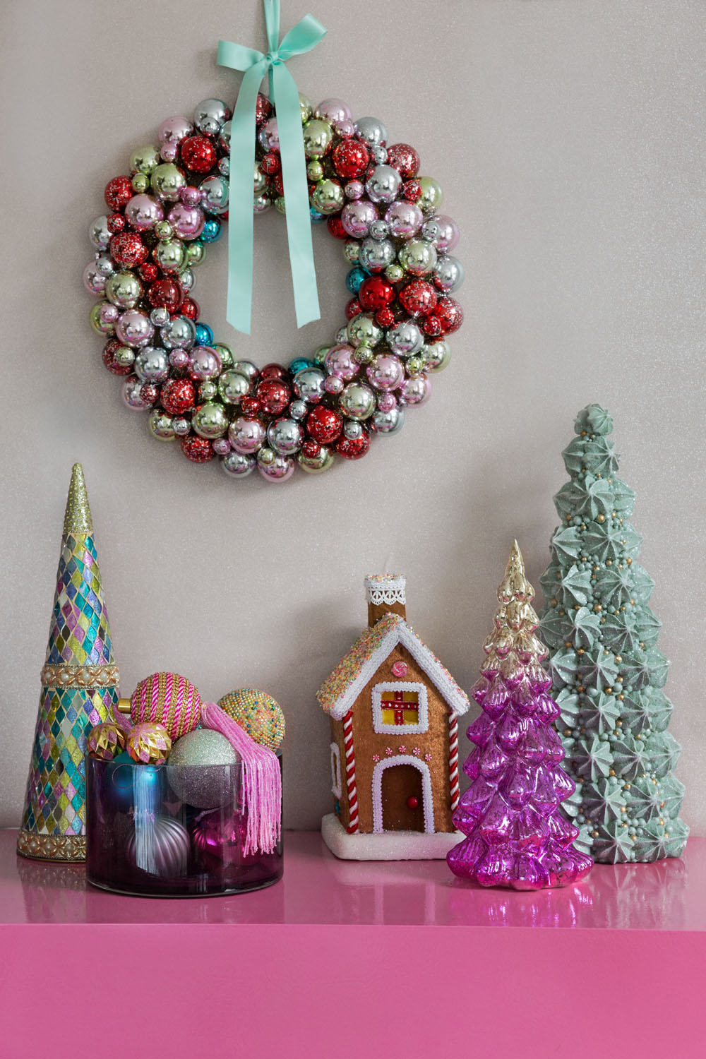 A festive wreath made of red, silver, and green baubles with a blue ribbon hangs on the wall above a pink table decorated with colourful cone-shaped trees, a gingerbread house, and a jar of baubles.