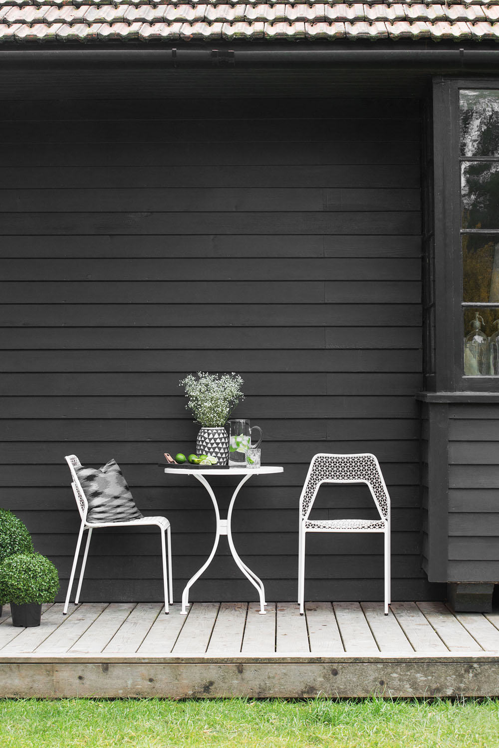 A small patio with two white metal chairs and a round table on a wooden decking outside a dark grey house. The table holds a vase with flowers, a jug, glasses, and a bowl of limes. One chair has a cushion.