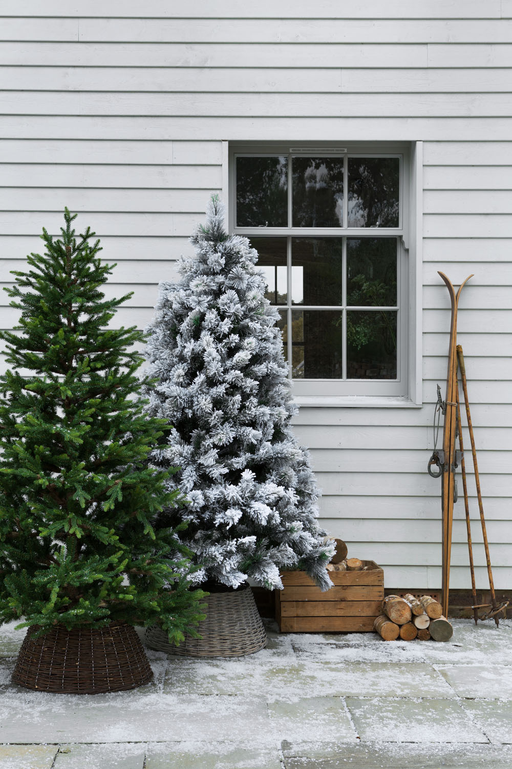 Two Christmas trees, one green and one covered in artificial snow, stand in baskets outside a white wood-panelled house with a window. Nearby are stacked firewood, a wooden crate, and a pair of vintage skis and poles.