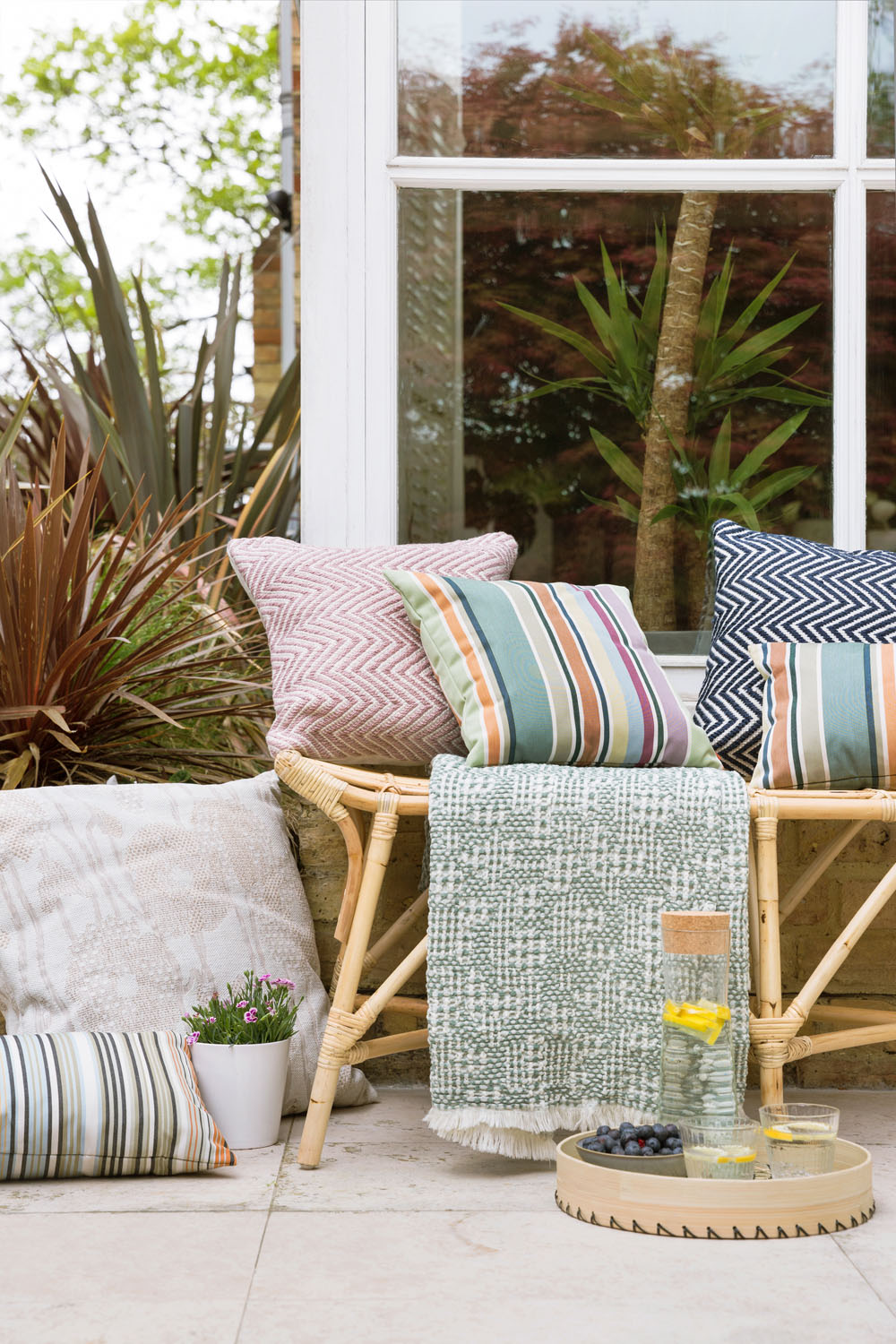 Outdoor seating area with a bamboo bench topped with patterned cushions and a green throw, next to potted plants. On the floor, there is a tray with a carafe of water, a glass, and a small bowl of berries.