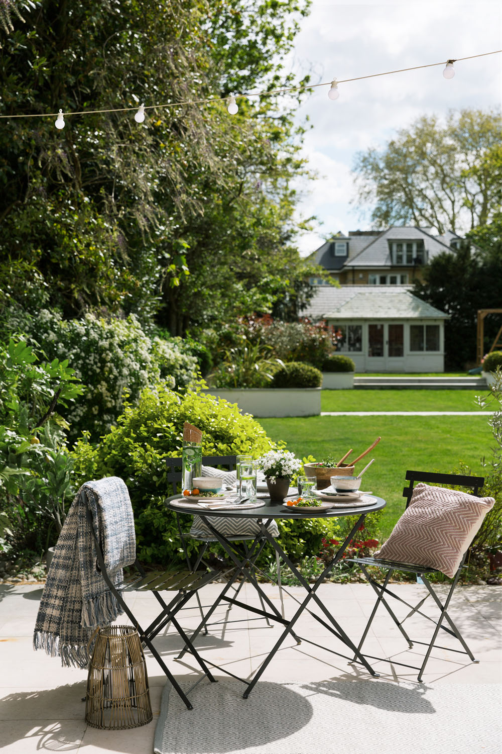 A small outdoor table with two chairs sits on a patio, set with plates, glasses, drinks, and food. A garden with green grass, trees, and a house in the background is visible under fairy lights.