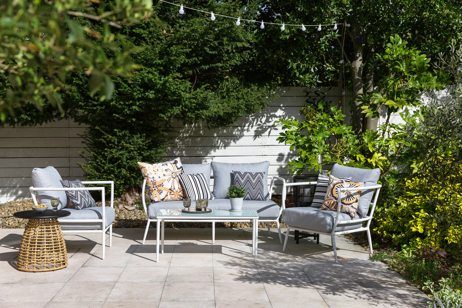 Outdoor patio with grey cushioned seating, patterned pillows, a glass-topped table with drinks, a wicker side table, potted plants, string lights overhead, and green trees and shrubs along a white fence in the background.