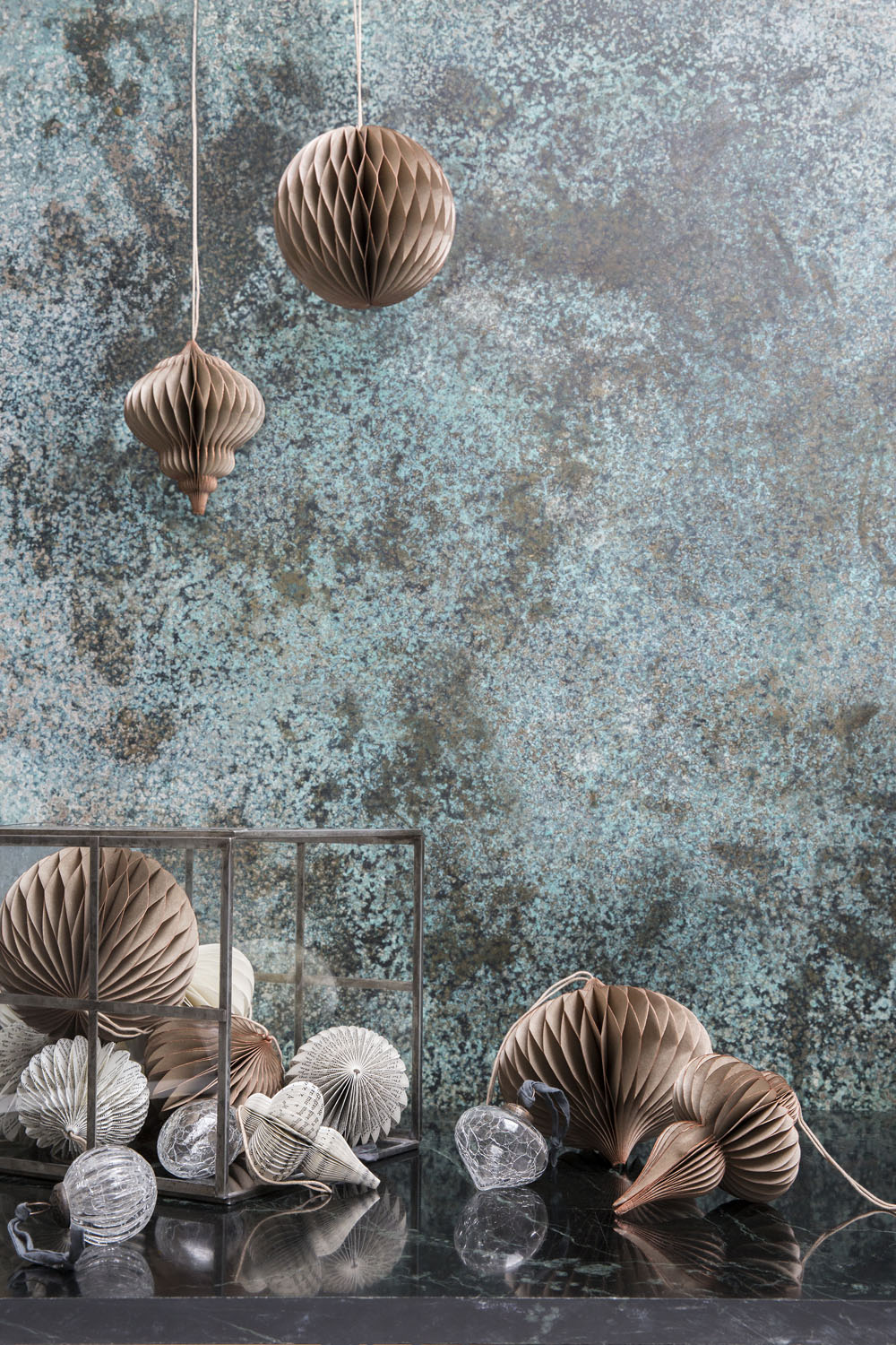 Brown and white paper ornaments are displayed, some hanging and some resting on a dark surface. Several ornaments are placed inside a glass box. The background features a textured teal and brown wall.