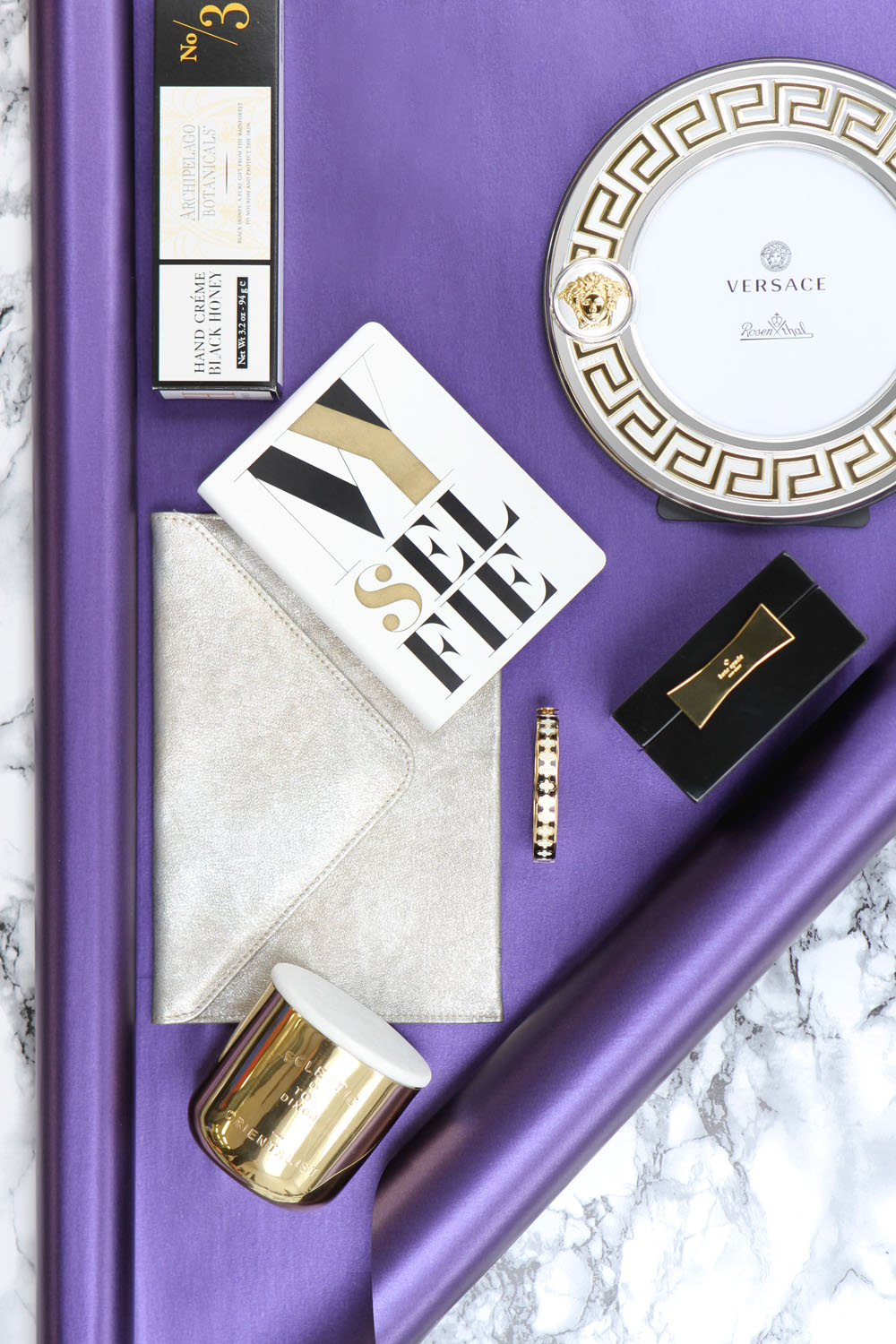 Flat lay of luxury items on a purple satin and marble background, including a Versace plate, gold candle, envelope clutch, SELFIE notebook, gold-patterned bracelet, lipstick, and boxed perfume.