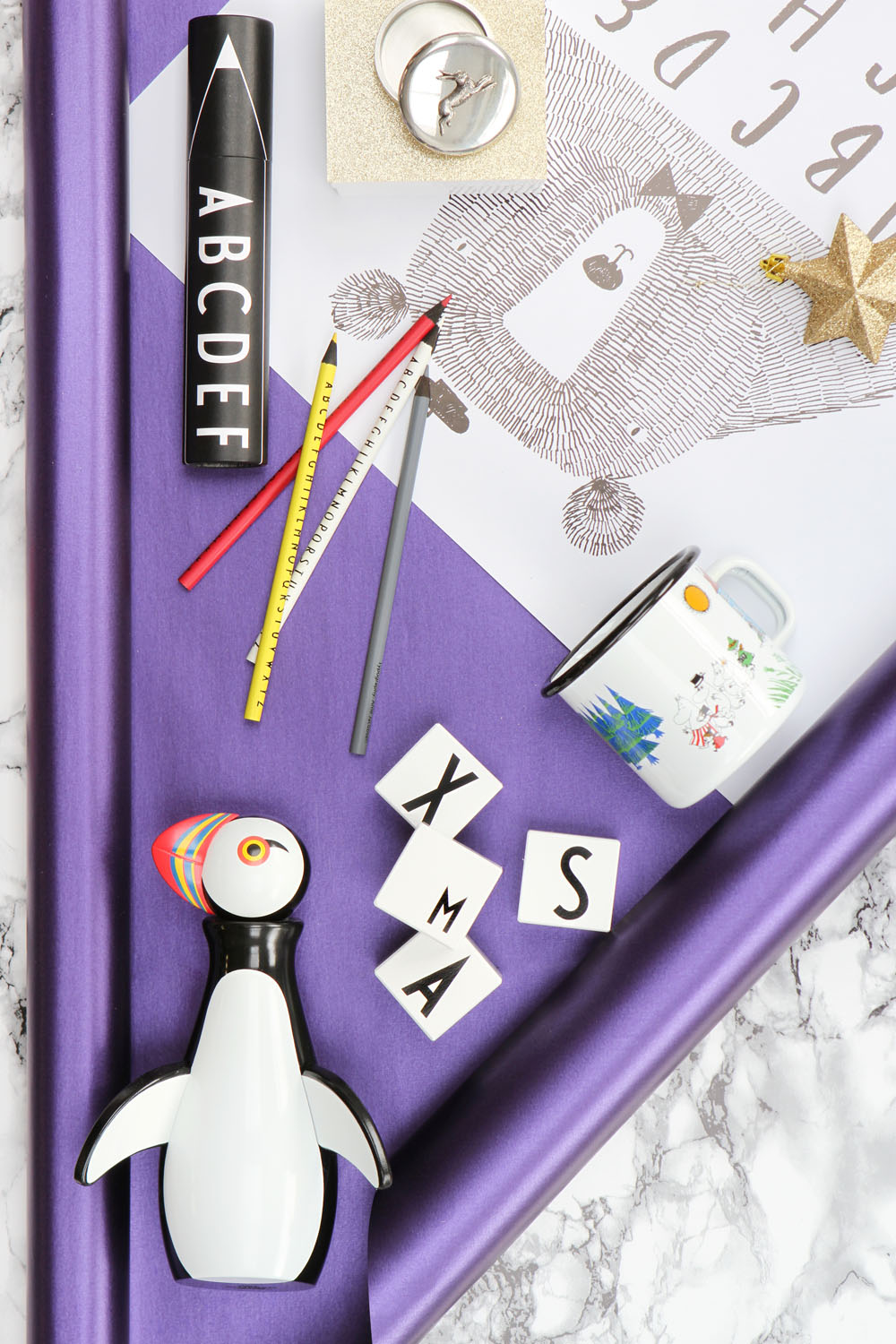 A flat lay features a black-and-white puffin figurine, pencils, rulers, letter tiles spelling XMAS, a mug, rings, a rolled paper tube labelled A-Z, and a print of a puffin, all on a purple satin surface.