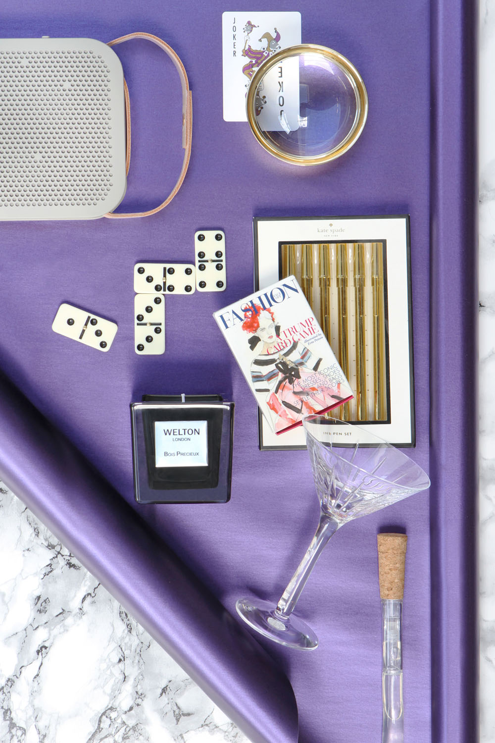 A flat lay on a purple background featuring a speaker, a joker playing card, a magnifying glass, dominoes, gold pens in a box, a fashion magazine, a black candle, a martini glass, and a glass vial.
