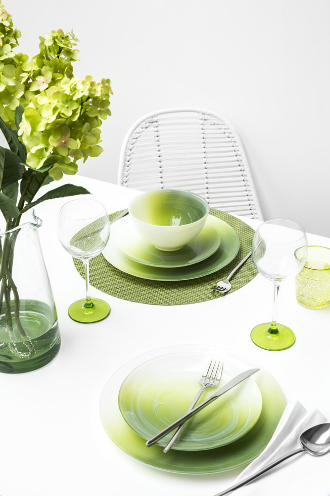 A white table set with green and white gradient plates, a matching bowl, a fork, knife, and spoon, two wine glasses with green bases, a textured green placemat, a glass, and a vase with green hydrangeas.