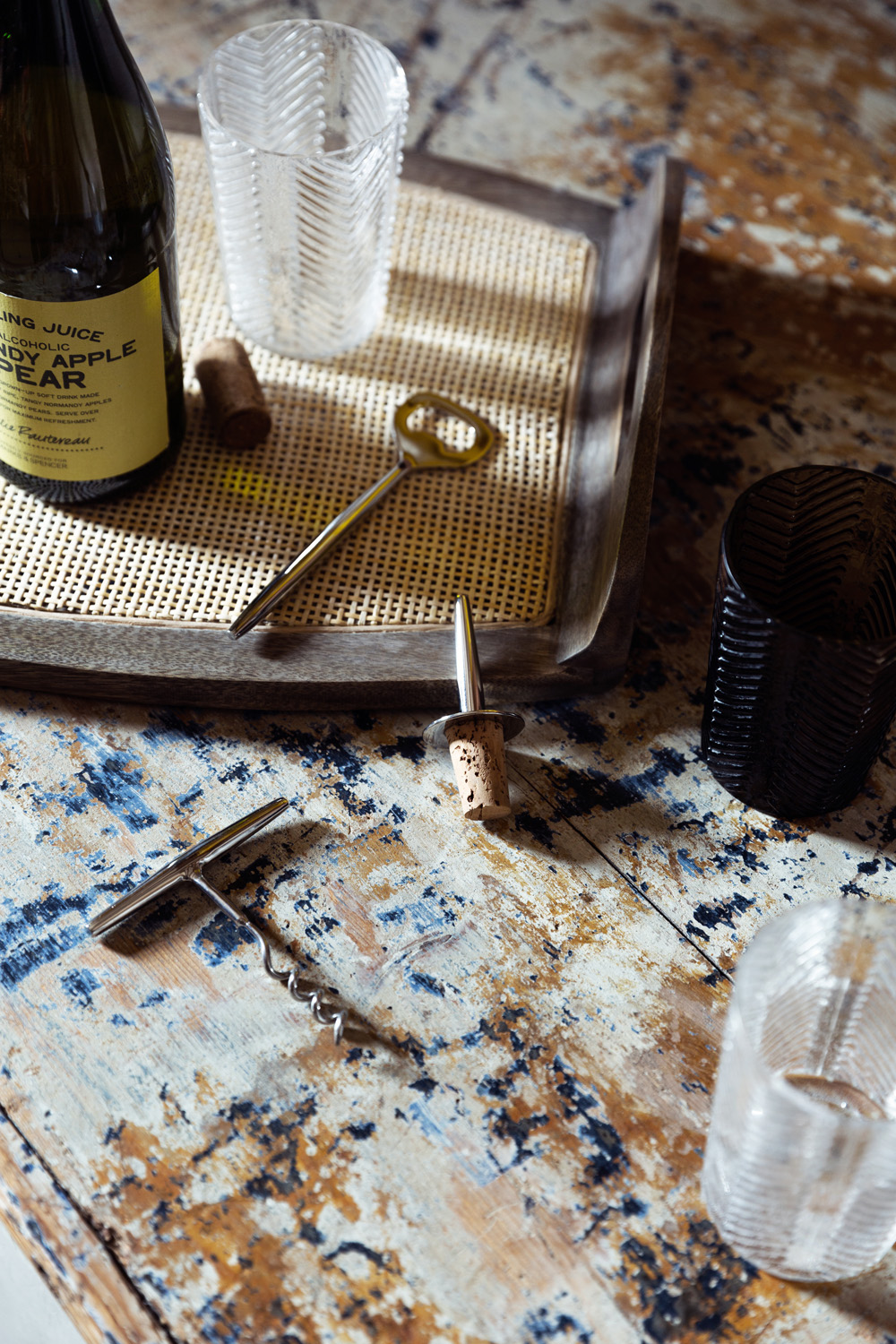 A bottle of sparkling juice, a cork, a corkscrew, a bottle stopper, and several textured glasses are arranged on a rustic, distressed wooden surface with a woven tray.