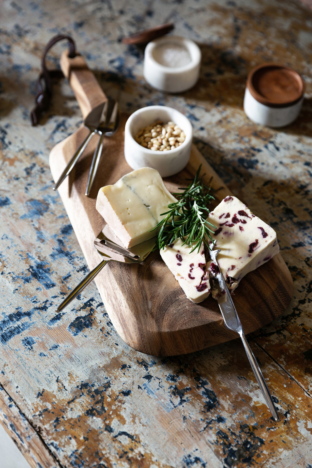 A wooden chopping board with two varieties of cheese, one with cranberries, garnished with rosemary and served with cheese knives. Small bowls with nuts and spices sit in the background on a rustic table.