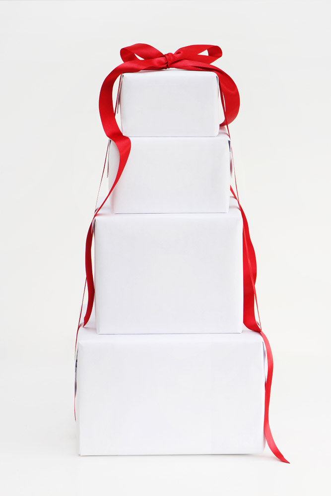 Four square boxes wrapped in plain white paper are stacked on top of each other in a pyramid shape. The stack is tied together with a long, red satin ribbon that is tied in a bow at the top.