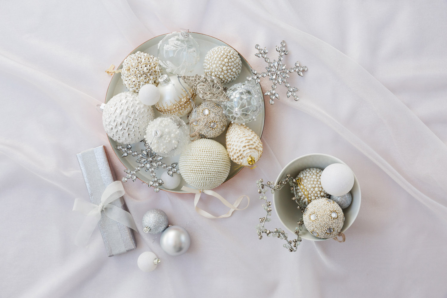 Two bowls are filled with white and silver Christmas baubles, including balls with beaded and textured surfaces. A wrapped silver present and silver snowflake decorations are nearby on a soft, pale fabric background.