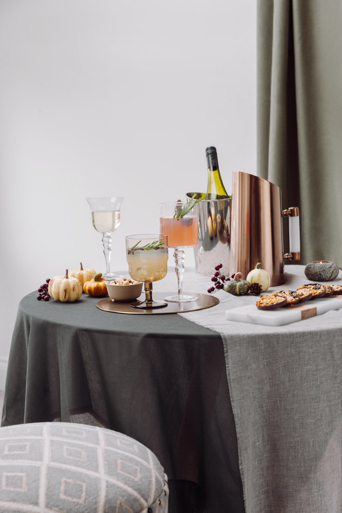 A round table with a grey tablecloth holds glasses with drinks, a bottle of wine, a copper ice bucket, small pumpkins, decorative gourds, grapes, and a patterned plate with snacks. A stool is in the foreground.