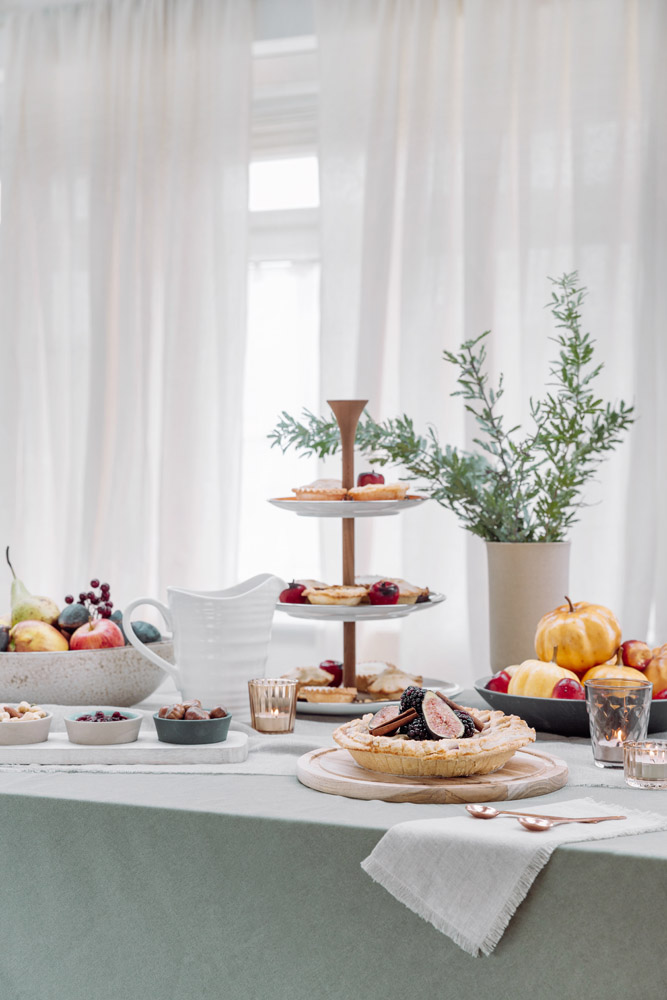 A table set with fruit, a tiered stand with pastries, a pie, bowls of nuts, and a jug. A vase with greenery and candles are also on the table. Sheer curtains let in light from the window behind.