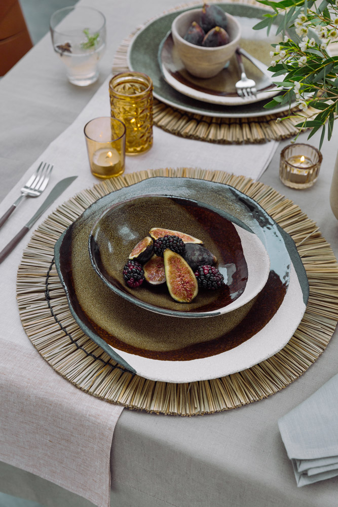 A table set with layered ceramic plates and bowls on a woven placemat, containing figs and blackberries. Amber and clear glass candle holders, cutlery, and a glass of water are arranged nearby on a light tablecloth.
