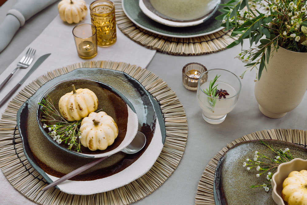 A table setting with rustic plates, small white pumpkins in bowls, cutlery, beige serviettes, candles in amber holders, a drink with star anise, and a ceramic vase with greenery and small white flowers.