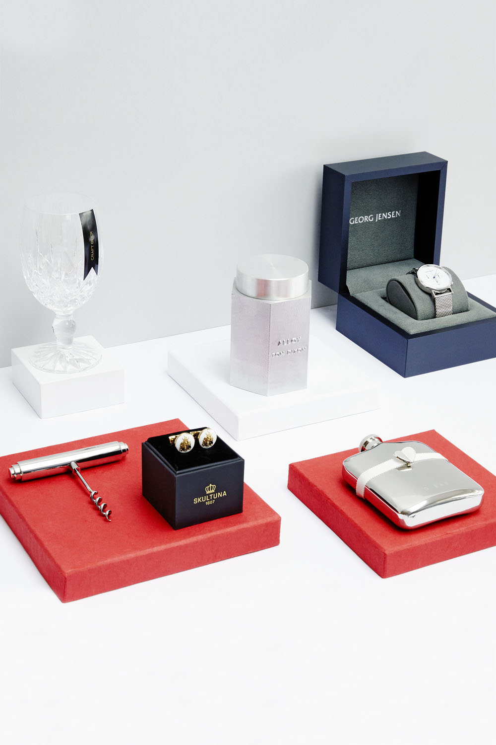 A selection of luxury gifts displayed on red and white plinths, including a crystal glass, metal hip flask, corkscrew, cufflinks, a silver container, and a wristwatch in a box.