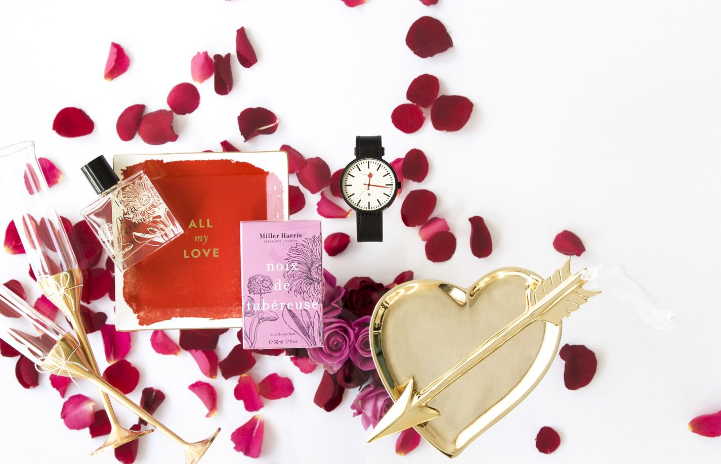 A flat lay of romantic-themed items, including rose petals, champagne glasses, a red card, a perfume bottle, a black-and-white wristwatch, and a gold heart-shaped tray with an arrow, on a white background.