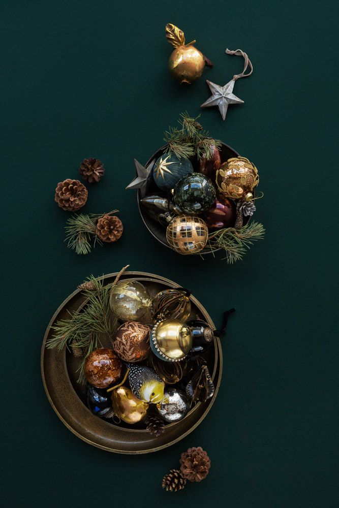 Two dark bowls filled with assorted Christmas baubles in gold, bronze, and dark green tones, surrounded by pine branches, pine cones, and a gold apple, arranged on a dark green background.