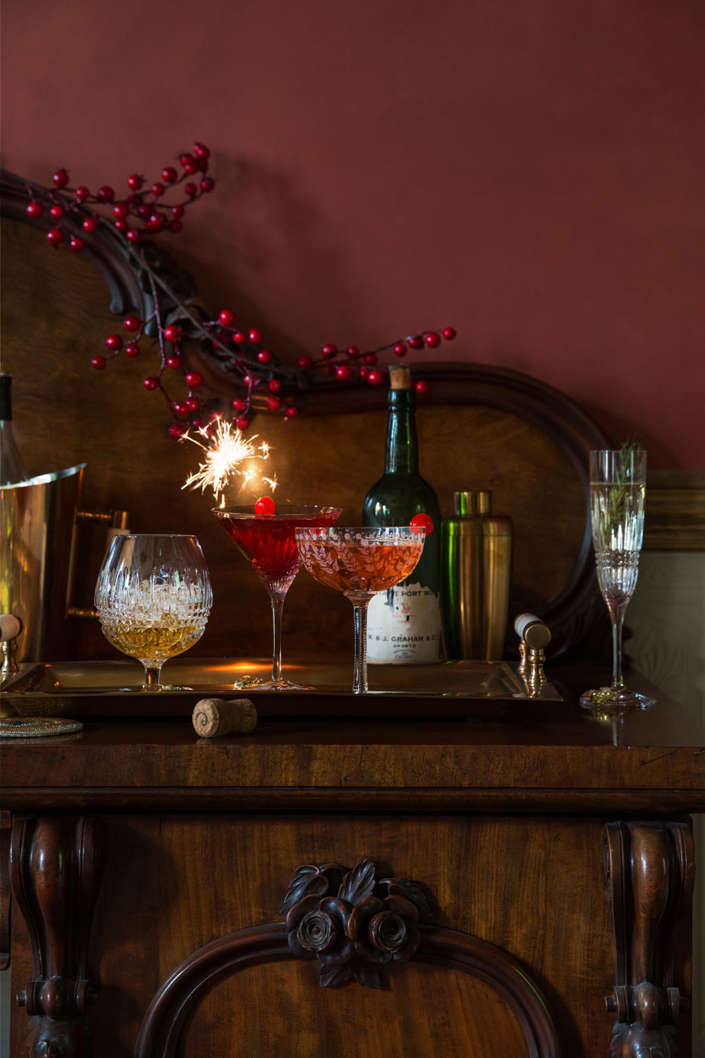 A wooden sideboard displays three crystal cocktail glasses, one with a sparkler, a wine bottle, a champagne flute, a metal ice bucket, a cocktail shaker, and a decorative branch with red berries.