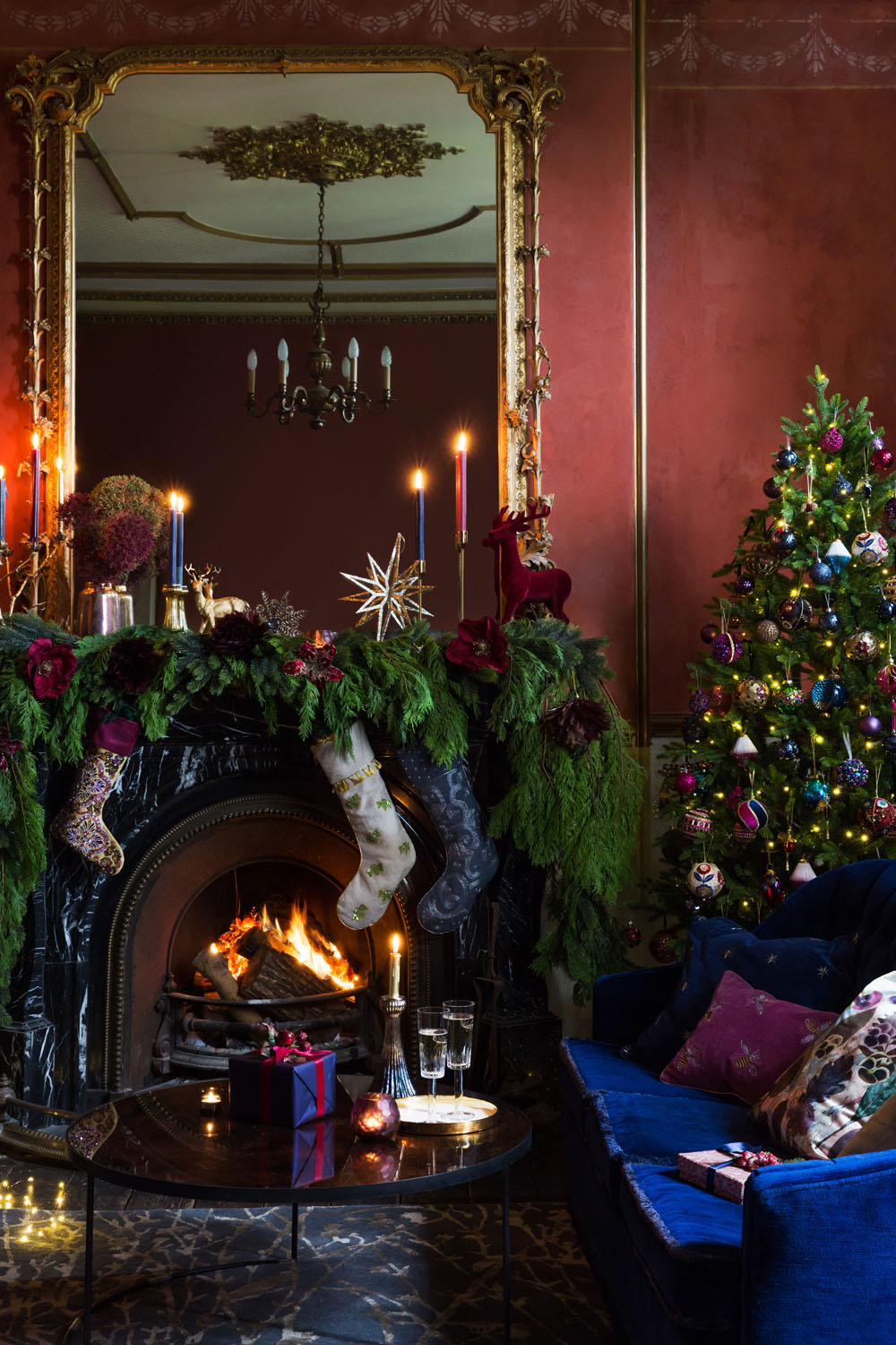 A decorated living room with a lit fireplace, stockings, candles, and festive garlands on the mantelpiece, a Christmas tree with baubles and fairy lights, a large mirror, and presents on a table in front of a blue sofa.