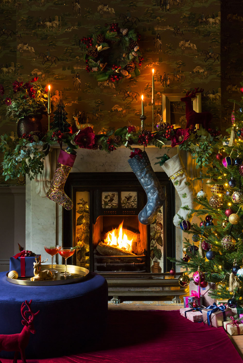 A decorated living room features a lit fireplace with three stockings hanging from the mantelpiece, a wreath above, a Christmas tree with baubles and presents, greenery, lit candles, and a table with drinks and a red reindeer figure.