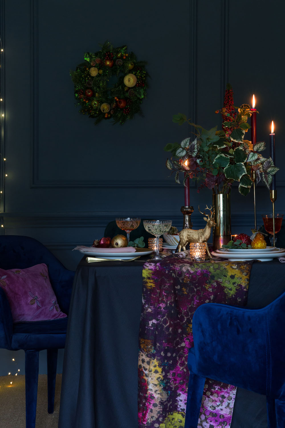 A dining table set with elegant glassware, a floral centrepiece, candles, and a decorative runner. Two blue velvet chairs are at the table. A wreath hangs on a dark blue wall with fairy lights in the background.